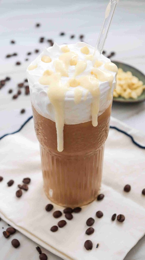 A glass of iced white chocolate mocha topped with whipped cream and white chocolate drizzle, served with a clear straw. Coffee beans are scattered on a white napkin, and a plate with white chocolate pieces is in the background.