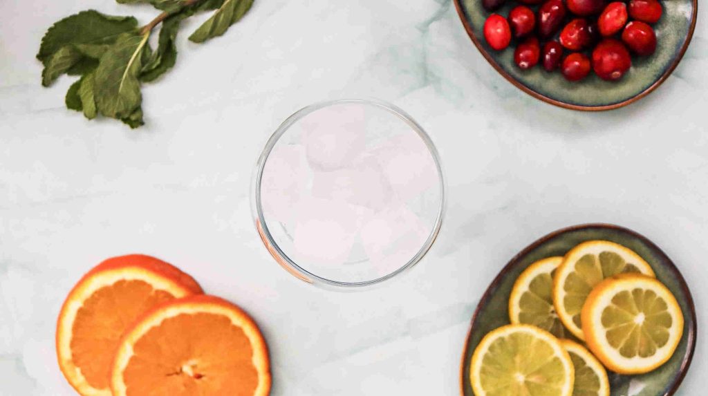 A glass of ice surrounded by orange slices, lemon slices, cranberries in a bowl, and fresh mint leaves on a light marble surface—perfect for crafting a refreshing cranberry juice mocktail.