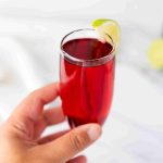 A hand holding a glass of dark red Mimosas with cranberry juice, garnished with a lime wedge on the rim, against a light, blurred background.