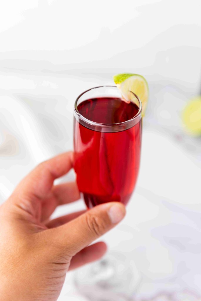 A hand holding a glass of dark red Mimosas with cranberry juice, garnished with a lime wedge on the rim, against a light, blurred background.