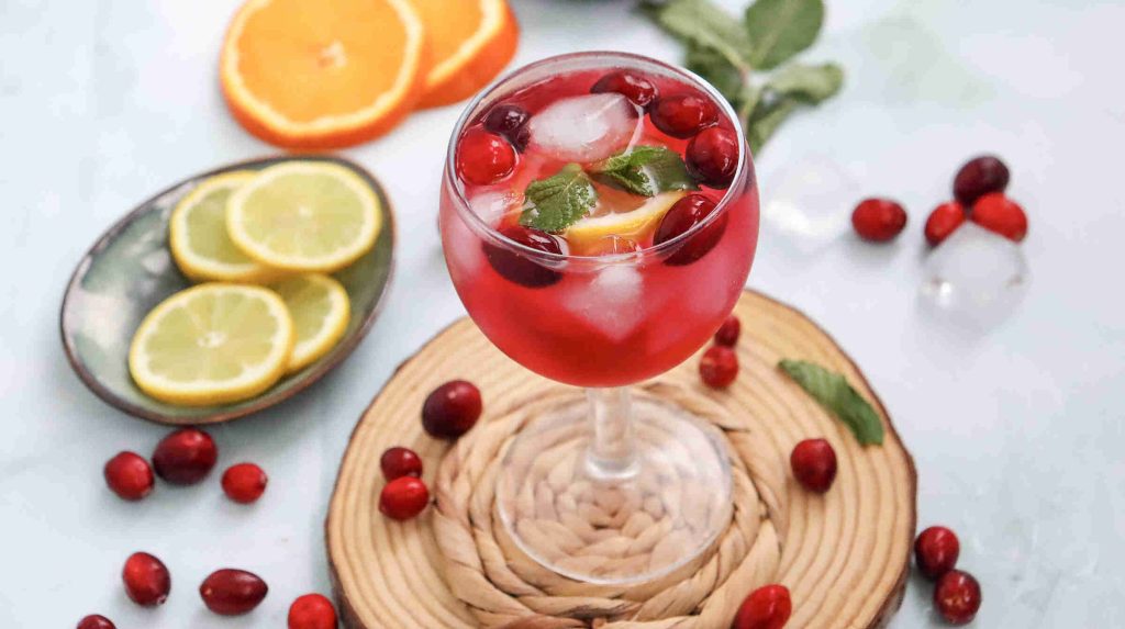 A glass of cranberry juice mocktail with ice, fresh cranberries, orange and lemon slices, and mint leaves sits on a woven coaster. Lemon and orange slices, cranberries, and mint are scattered around on a light surface.