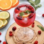 A glass of cranberry juice mocktail with ice, fresh cranberries, orange and lemon slices, and mint leaves sits on a woven coaster. Lemon and orange slices, cranberries, and mint are scattered around on a light surface.