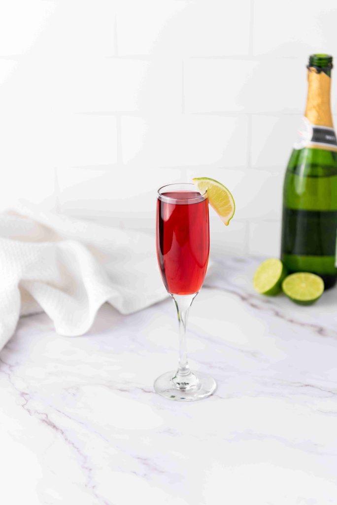 A flute glass filled with a red cocktail, inspired by mimosas with cranberry juice, garnished with a lime wedge and set on a marble surface. In the background, a green champagne bottle and sliced lime are visible.