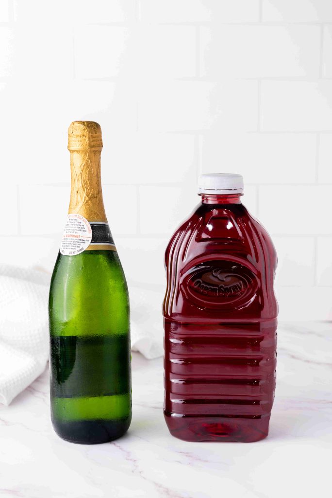 A green bottle of champagne with a gold foil top stands next to a large, clear plastic bottle of cranberry juice—perfect for making mimosas with cranberry juice—on a white marble surface with a white tile background.
