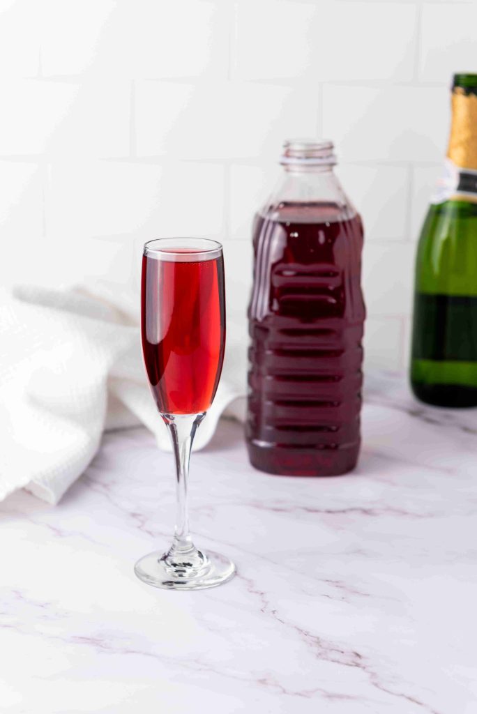 A glass of red liquid, reminiscent of mimosas with cranberry juice, stands on a marble surface. In the background are a plastic bottle of the same liquid and a green glass bottle, with a white cloth off to the side.