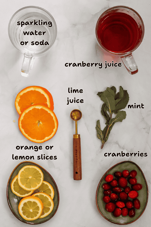 A flat lay of ingredients for a cranberry juice mocktail: a glass of sparkling water, cranberry juice, orange slices, a bowl of cranberries, a mint sprig, and a metal spoon with lime juice on a marble surface.