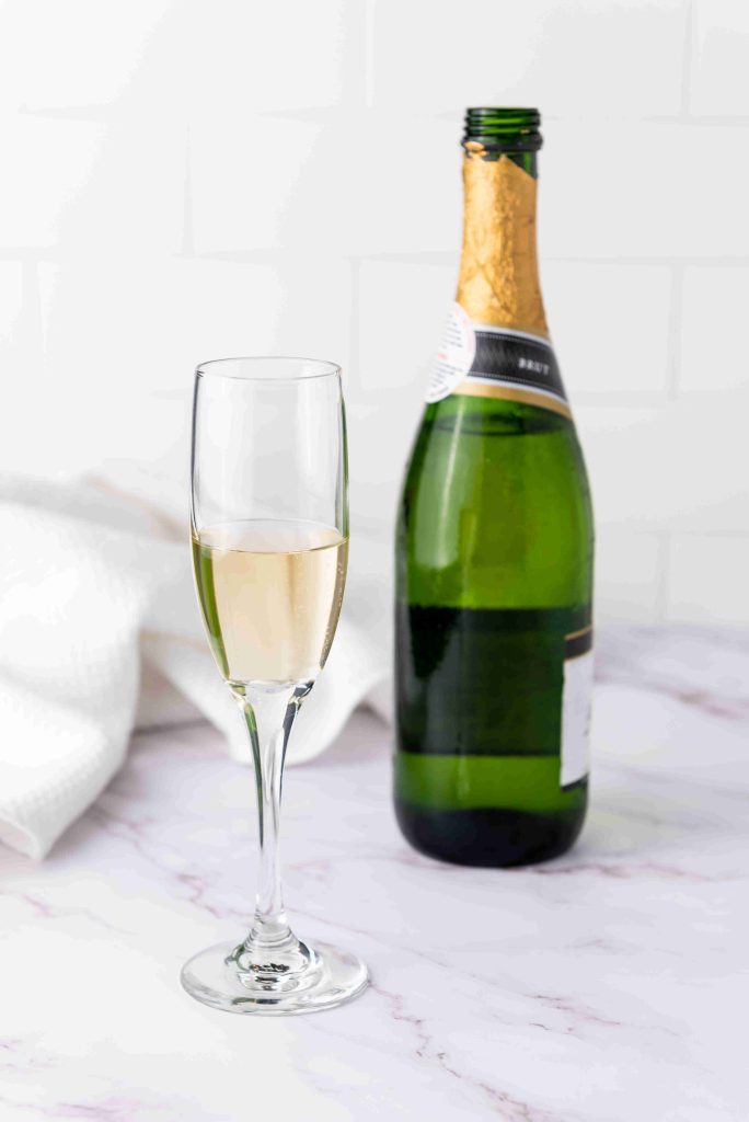A glass of champagne, perfect for mimosas with cranberry juice, sits beside an open green bottle on a marble surface, with a white towel in the background.