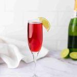A flute glass filled with a red drink, garnished with a lime wedge on the rim. In the background, Mimosas with cranberry juice are set alongside a champagne bottle, lime halves, and a white cloth on a marble surface.