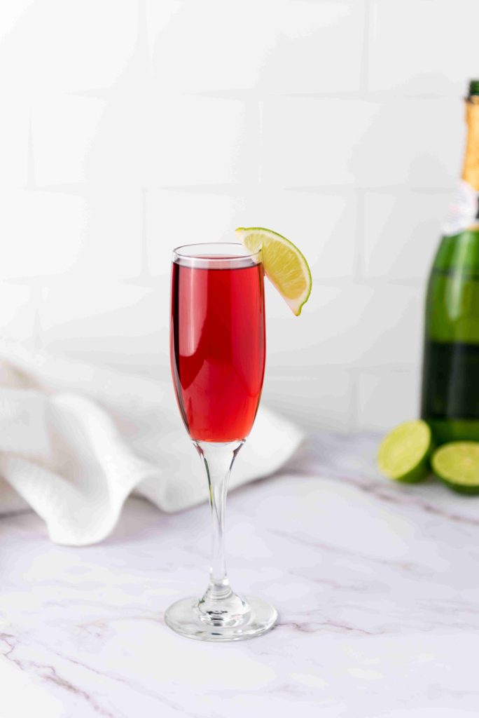 A flute glass filled with a red drink, garnished with a lime wedge on the rim. In the background, Mimosas with cranberry juice are set alongside a champagne bottle, lime halves, and a white cloth on a marble surface.