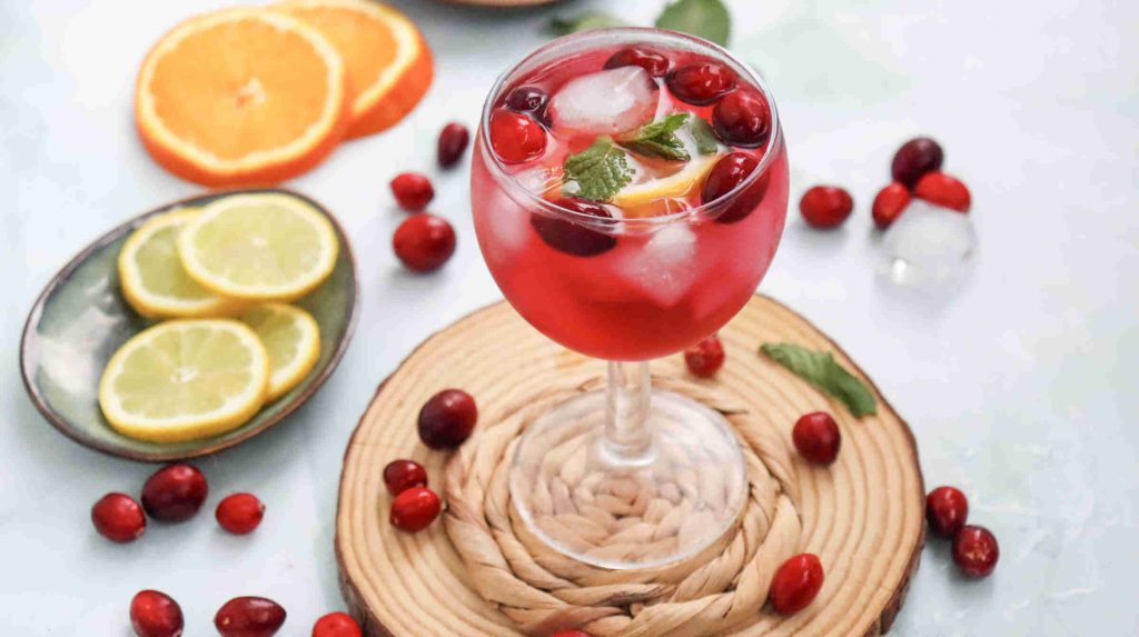A glass of cranberry juice mocktail with ice, fresh cranberries, a lemon slice, and mint garnish sits on a woven mat. Orange and lemon slices and scattered cranberries surround the glass on a light surface.