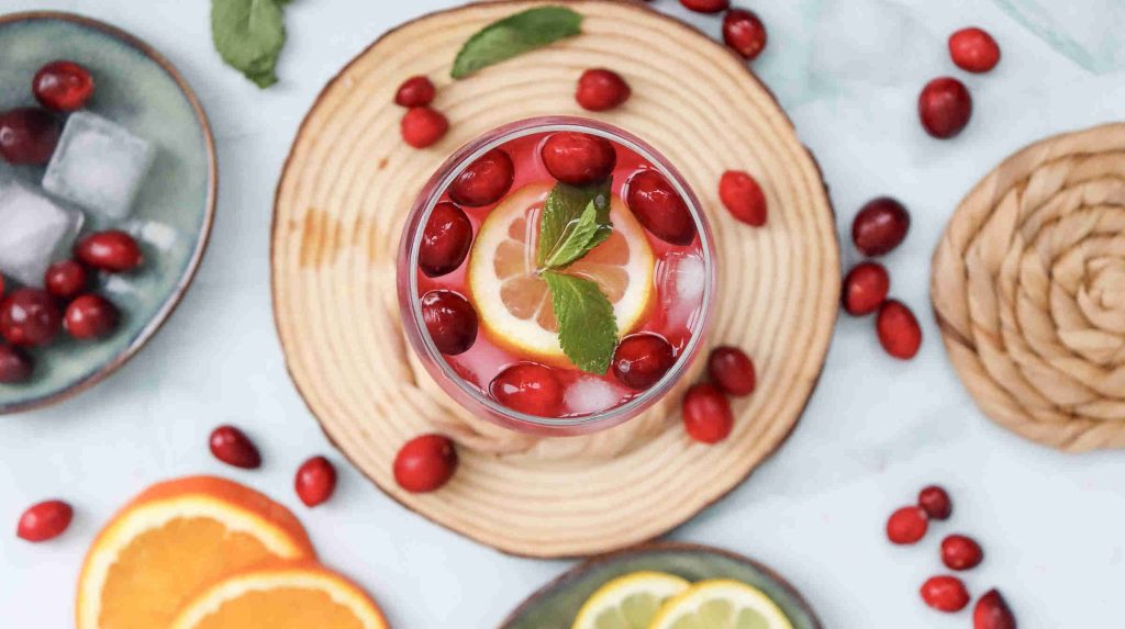 A top-down view of a cranberry juice mocktail with lemon slices, cranberries, and mint, surrounded by scattered cranberries and citrus slices on a white surface.