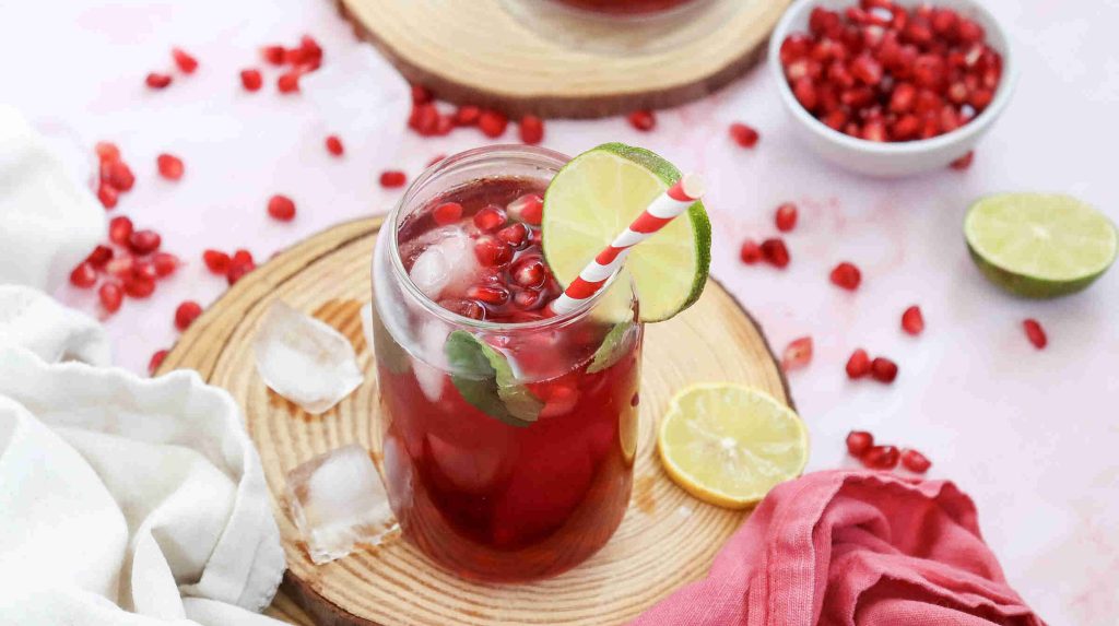 A vibrant Mocktail with Pomegranate Juice Recipe shines in a glass with ice, mint, lime slice, and a red-striped straw on a wooden coaster. Pomegranate seeds, lime halves, and scattered ice cubes complete the refreshing scene.