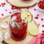 A vibrant Mocktail with Pomegranate Juice Recipe shines in a glass with ice, mint, lime slice, and a red-striped straw on a wooden coaster. Pomegranate seeds, lime halves, and scattered ice cubes complete the refreshing scene.