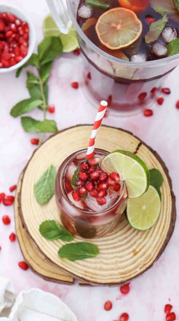 A glass of iced mocktail with pomegranate juice recipe, garnished with lime slices, mint leaves, and pomegranate seeds, sits with a red-striped straw on a wooden coaster. A pitcher and fresh ingredients appear in the background.