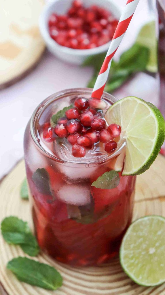 A glass of iced pomegranate drink, made using a refreshing Mocktail with Pomegranate Juice Recipe, is garnished with fresh pomegranate seeds, mint leaves, and a lime wheel. A red-and-white striped straw, halved lime, and more mint leaves are nearby.