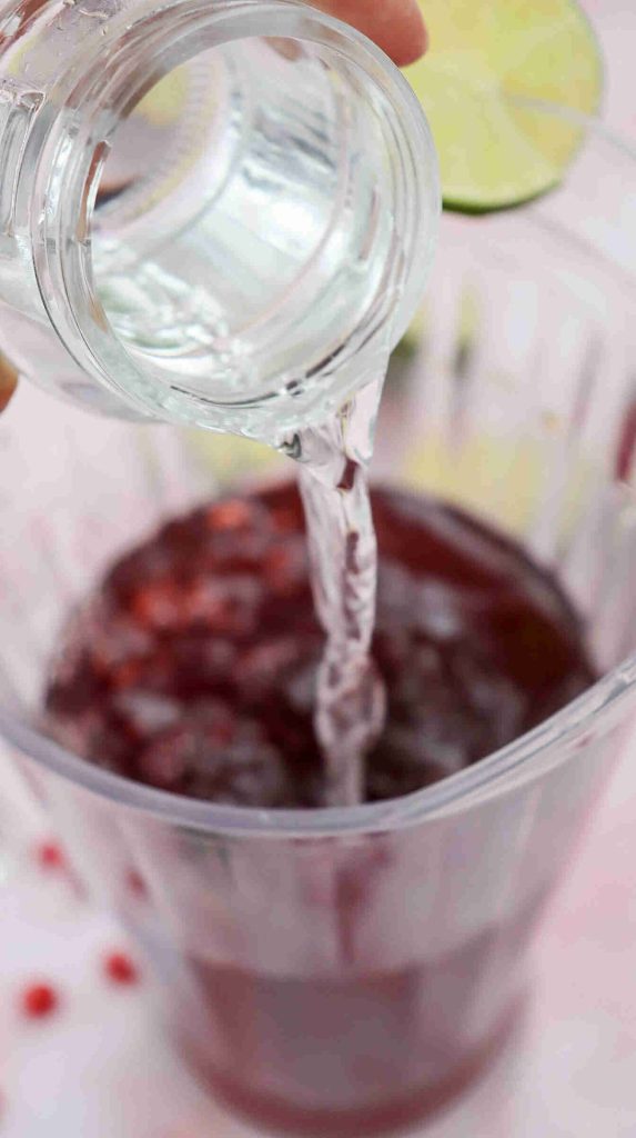 A clear liquid is being poured from a small glass bottle into a plastic cup filled with a dark red beverage, ice, and a slice of lime on the rim—a refreshing Mocktail with Pomegranate Juice Recipe in the making.