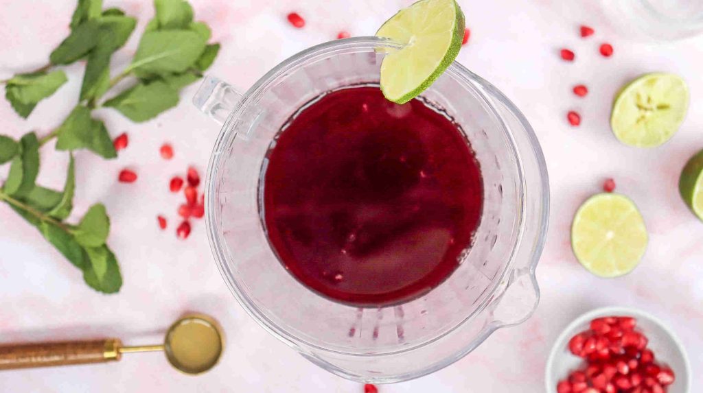A glass pitcher filled with dark red juice, garnished with a lime slice. Around it are fresh mint leaves, lime halves, pomegranate seeds, a gold spoon, and a bowl of pomegranate seeds—perfect for a Mocktail with Pomegranate Juice Recipe.