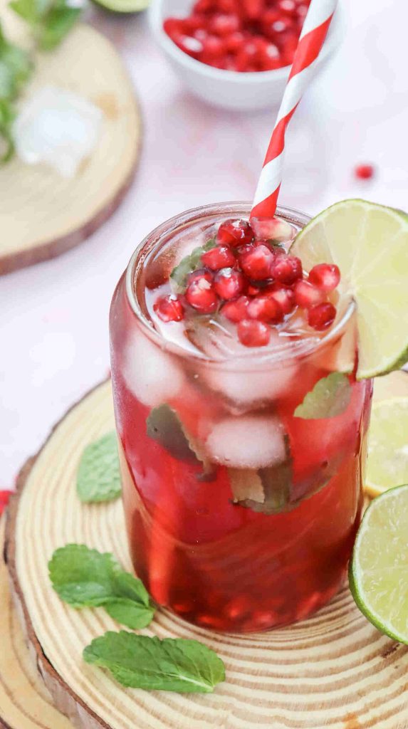 A refreshing glass of Mocktail with Pomegranate Juice Recipe, garnished with pomegranate seeds, lime slices, mint leaves, and a striped straw, sits on a wooden coaster with extra mint and lime on the side.