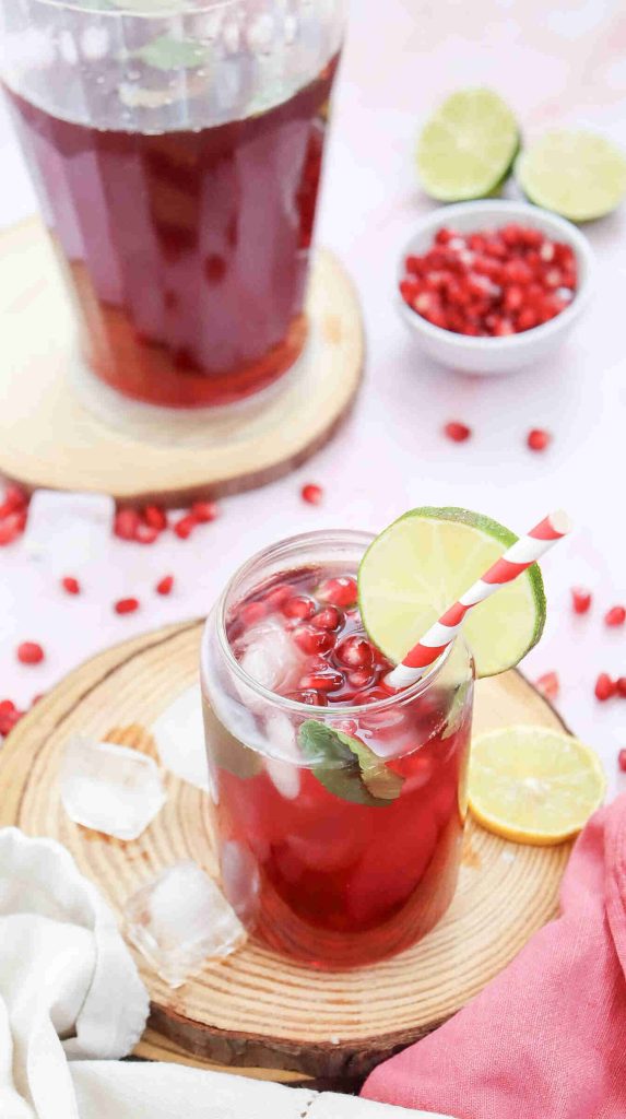 A refreshing Mocktail with Pomegranate Juice Recipe: a glass filled with ice, pomegranate seeds, mint leaves, and lime slice, served with a red-and-white striped straw. A jug of juice, lime slices, and pomegranate seeds sit in the background.