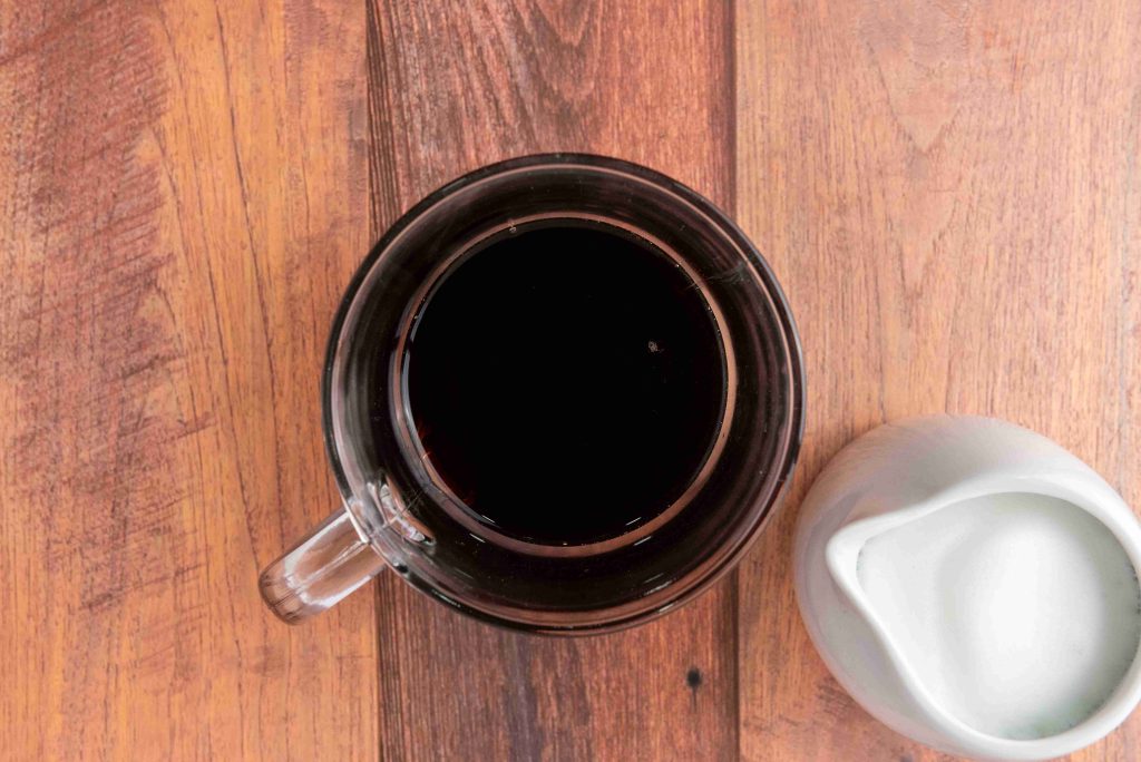 A glass cup filled with black coffee sits on a wooden surface next to a white ceramic creamer filled with milk—perfect for cozying up with your favorite Christmas coffee or trying a new gingerbread latte recipe.