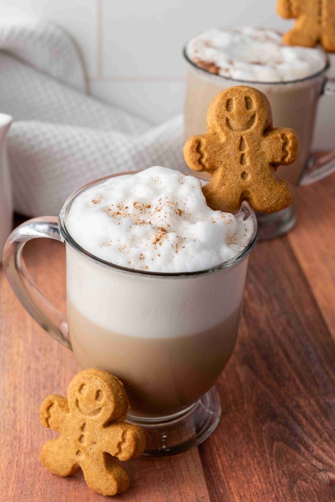 A glass mug of frothy latte topped with cinnamon and a smiling gingerbread cookie, with another mug and cookie in the background on a wooden surface—a perfect Christmas coffee inspired by a classic gingerbread latte recipe.