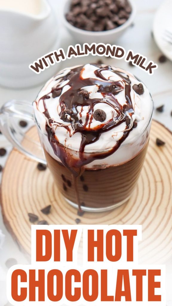 A glass mug of hot chocolate topped with whipped cream, chocolate syrup, and chocolate chips sits on a wooden coaster. Perfect for drinks for the fall, this DIY treat uses almond milk and features chocolate chips in the background.