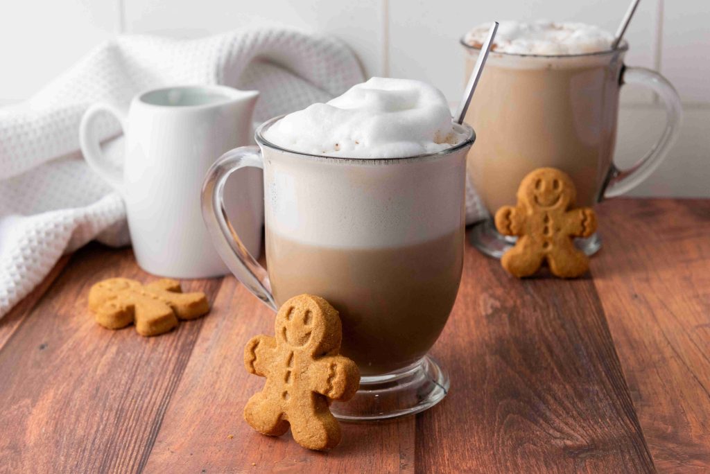 Two glass mugs of Christmas coffee topped with foam sit on a wooden table, each with a metal spoon. Smiling gingerbread cookies are placed beside the mugs, and a white creamer is in the background.