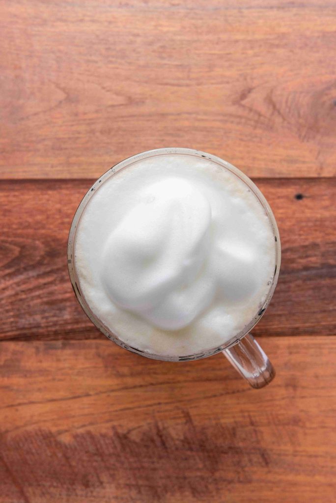 A glass cup filled with frothy, foamy milk sits on a wooden surface, viewed from above—perfect for a gingerbread latte recipe or your favorite Christmas coffee.