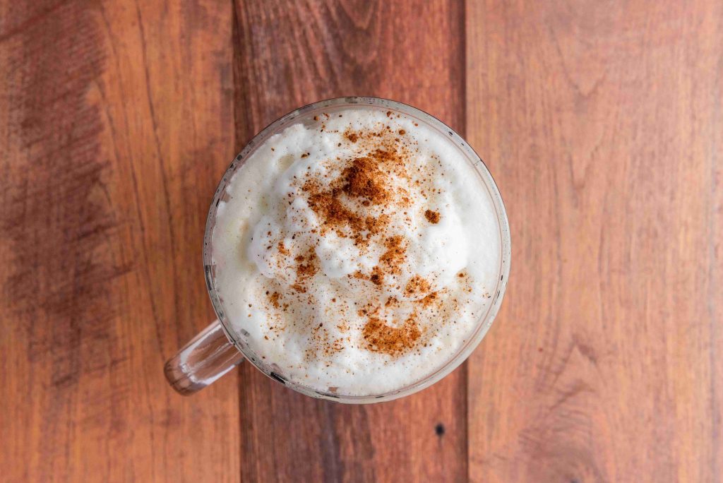 A clear glass mug filled with a creamy, foamy Christmas coffee topped with a sprinkle of cinnamon, sitting on a wooden surface.