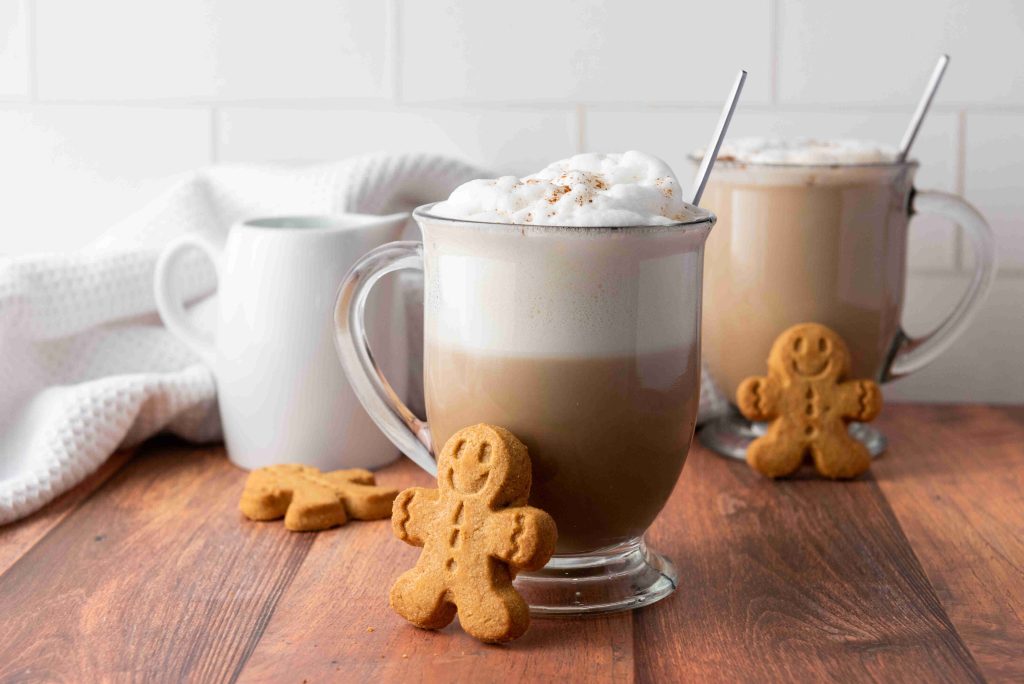 Two glass mugs of frothy coffee sit on a wooden table, each with a metal spoon and a gingerbread cookie—perfect for trying your favorite Christmas coffee or testing out a new gingerbread latte recipe. A white pitcher and towel appear in the background.