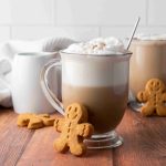 Two glass mugs of frothy coffee sit on a wooden table, each with a metal spoon and a gingerbread cookie—perfect for trying your favorite Christmas coffee or testing out a new gingerbread latte recipe. A white pitcher and towel appear in the background.