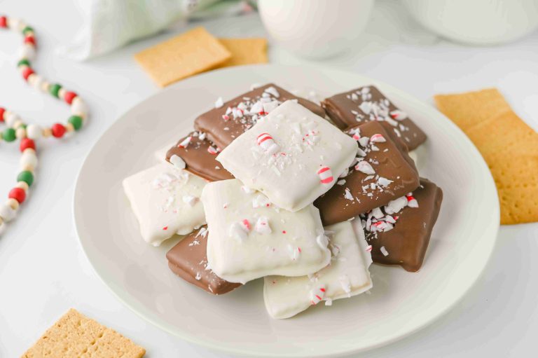 A plate of holiday chocolate covered graham crackers—square treats coated in white and milk chocolate, topped with crushed peppermint candy—sits next to whole graham crackers and a colorful candy necklace.