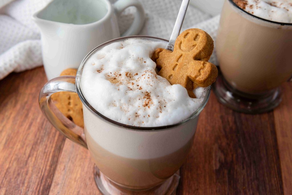 A glass mug of frothy latte topped with cinnamon and a gingerbread cookie, with a straw, sits on a wooden table—perfect inspiration for your next Christmas coffee or homemade gingerbread latte recipe. Another mug and small white pitcher are in the background.