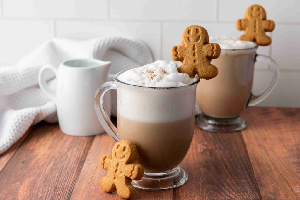 Two glass mugs of frothy Christmas coffee topped with foam and a sprinkle of cinnamon, each garnished with a gingerbread cookie. A white pitcher and a textured cloth are in the background on a wooden table.