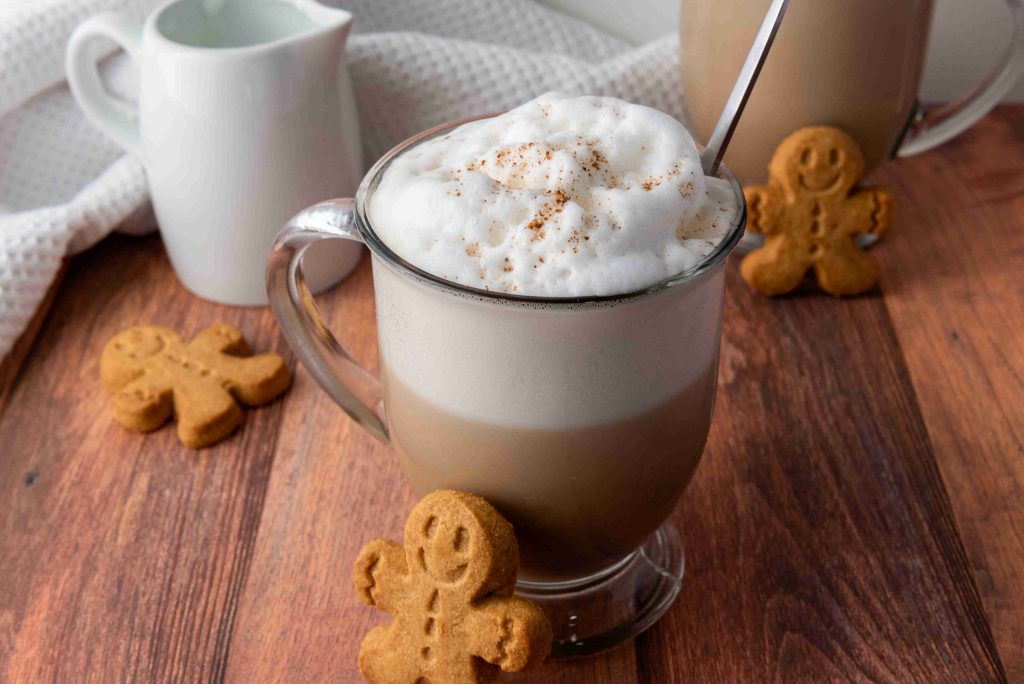 A glass mug of frothy coffee sits on a wooden table, topped with foam and sprinkled with cinnamon. Gingerbread cookies shaped like people surround this Christmas coffee, with a white creamer and cloth in the background.