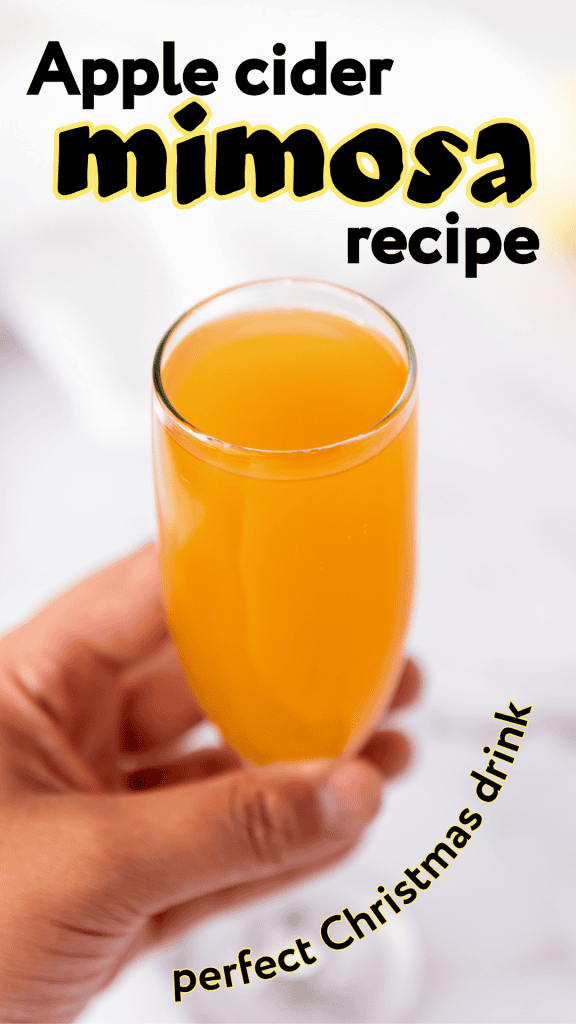 A hand holding a champagne glass filled with orange apple cider mimosa. Text on the image reads: “Apple cider mimosa recipe, the perfect sparkling holiday mimosa for Christmas.”.