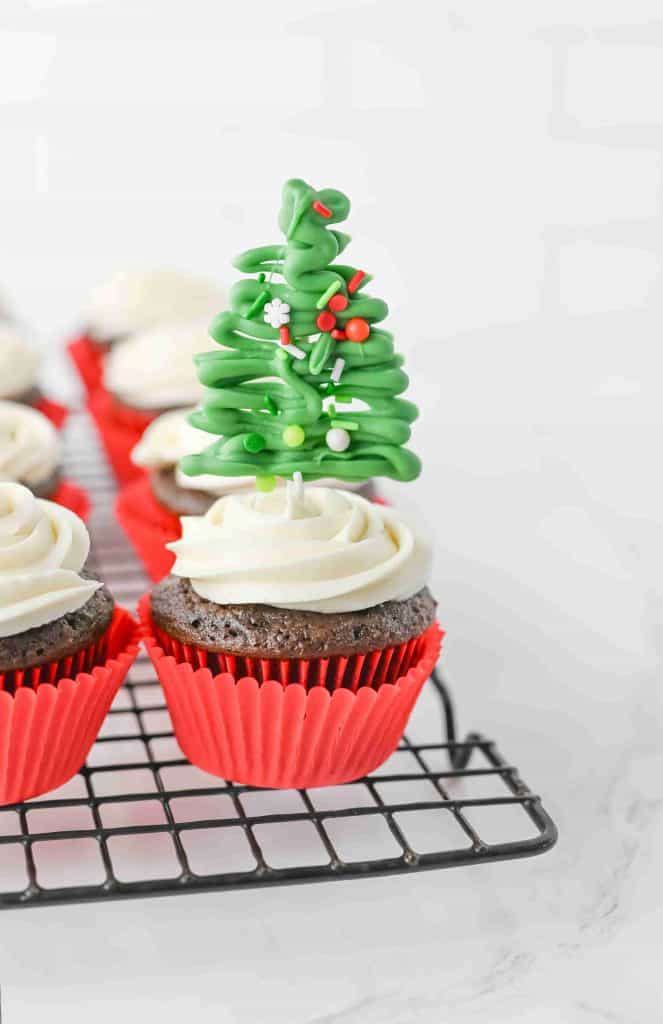 Christmas tree cupcakes: Chocolate cupcakes with white frosting in red wrappers on a cooling rack; one cupcake is topped with a decorative green Christmas tree made of icing and sprinkles.