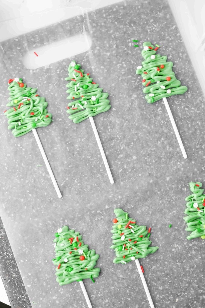 Six christmas tree cupcakes made of green icing on white sticks are arranged on a gray cutting board. Each tree-shaped treat is decorated with festive red, white, and green sprinkles.