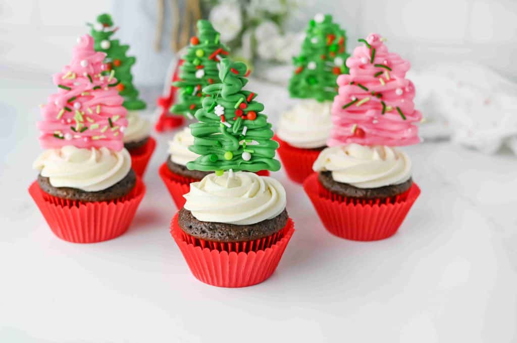 Christmas tree cupcakes: Chocolate cupcakes with white frosting in red wrappers, each topped with a decorative Christmas tree made of green or pink icing and sprinkles. The background is white with blurred flowers.
