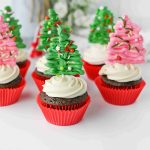 Christmas tree cupcakes: Chocolate cupcakes with white frosting in red wrappers, each topped with a decorative Christmas tree made of green or pink icing and sprinkles. The background is white with blurred flowers.
