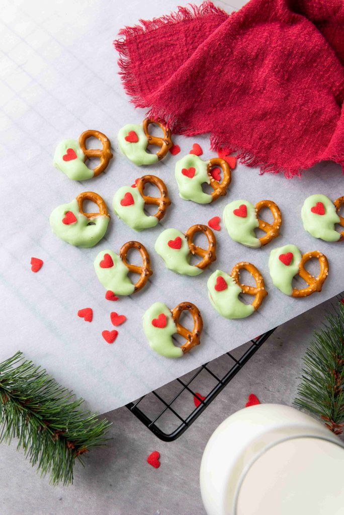 A cooling rack holds Grinch Heart Pretzels—dipped-pretzels-for-christmas in green coating and topped with red heart sprinkles. Nearby, a glass of milk, red cloth, and pine branches rest on a light surface.