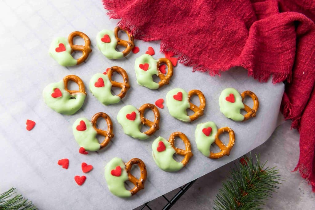 Grinch Heart Pretzels dipped in green coating with red heart sprinkles are arranged on parchment paper next to a red cloth and pine branch, making these dipped-pretzels-for-christmas the perfect festive, holiday-themed snack.