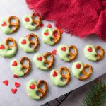Grinch Heart Pretzels dipped in green coating with red heart sprinkles are arranged on parchment paper next to a red cloth and pine branch, making these dipped-pretzels-for-christmas the perfect festive, holiday-themed snack.