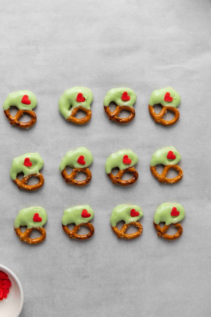 Twelve Grinch Heart Pretzels are half-dipped in green coating and topped with small red heart decorations, arranged neatly on a gray surface. A bowl with red heart candies completes this festive dipped-pretzels-for-christmas display.