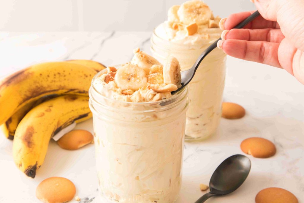 Two mason jars filled with Christmas Banana Pudding, topped with banana slices and vanilla wafers. A hand holds a spoon in one jar. Bananas and more wafers are on a white marble surface nearby.