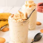 Two mason jars filled with Christmas Banana Pudding, topped with banana slices and vanilla wafers. A hand holds a spoon in one jar. Bananas and more wafers are on a white marble surface nearby.