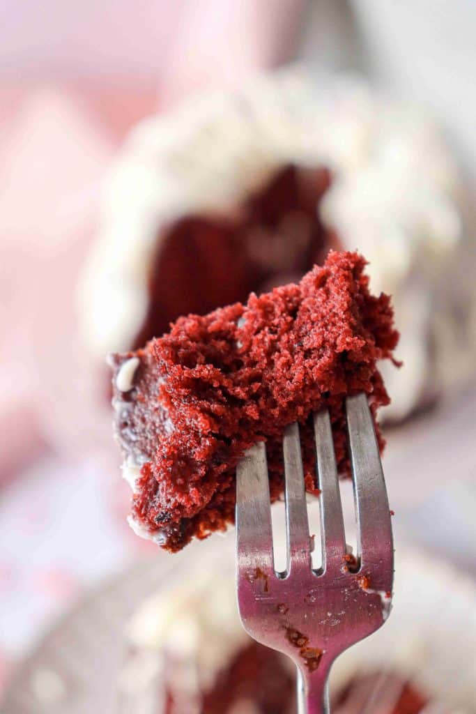 A close-up of a fork holding a bite of moist Christmas Red Velvet Bundt Cake with white frosting, with more cake and a blurred background visible behind.