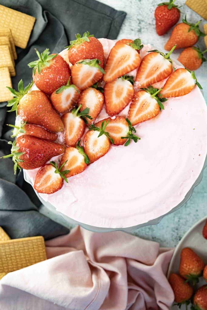 A round Christmas strawberry cheesecake with pink frosting is topped with neat rows of fresh halved strawberries. The cake is surrounded by a gray and a pink cloth, with whole strawberries and rectangular cookies nearby.