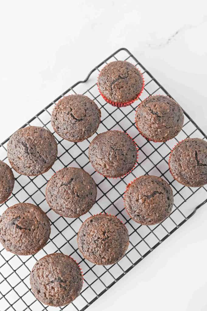 Twelve chocolate muffins with red paper liners, resembling festive christmas tree cupcakes, cooling on a black wire rack against a white background.