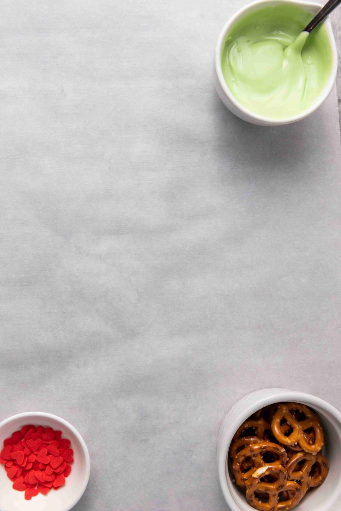 Three bowls on a parchment surface: one with green melted chocolate and a spoon (top right), one with red heart sprinkles (bottom left), and one with pretzels (bottom right)—perfect for making Grinch Heart Pretzels or dipped-pretzels-for-christmas.
